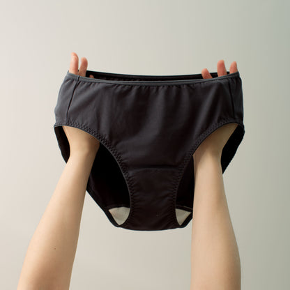 Hands holding up a Hold Panty, a black mid-waist brief panty with a specially constructed white gusset designed to work with The Stay Liner reusable panty liner.