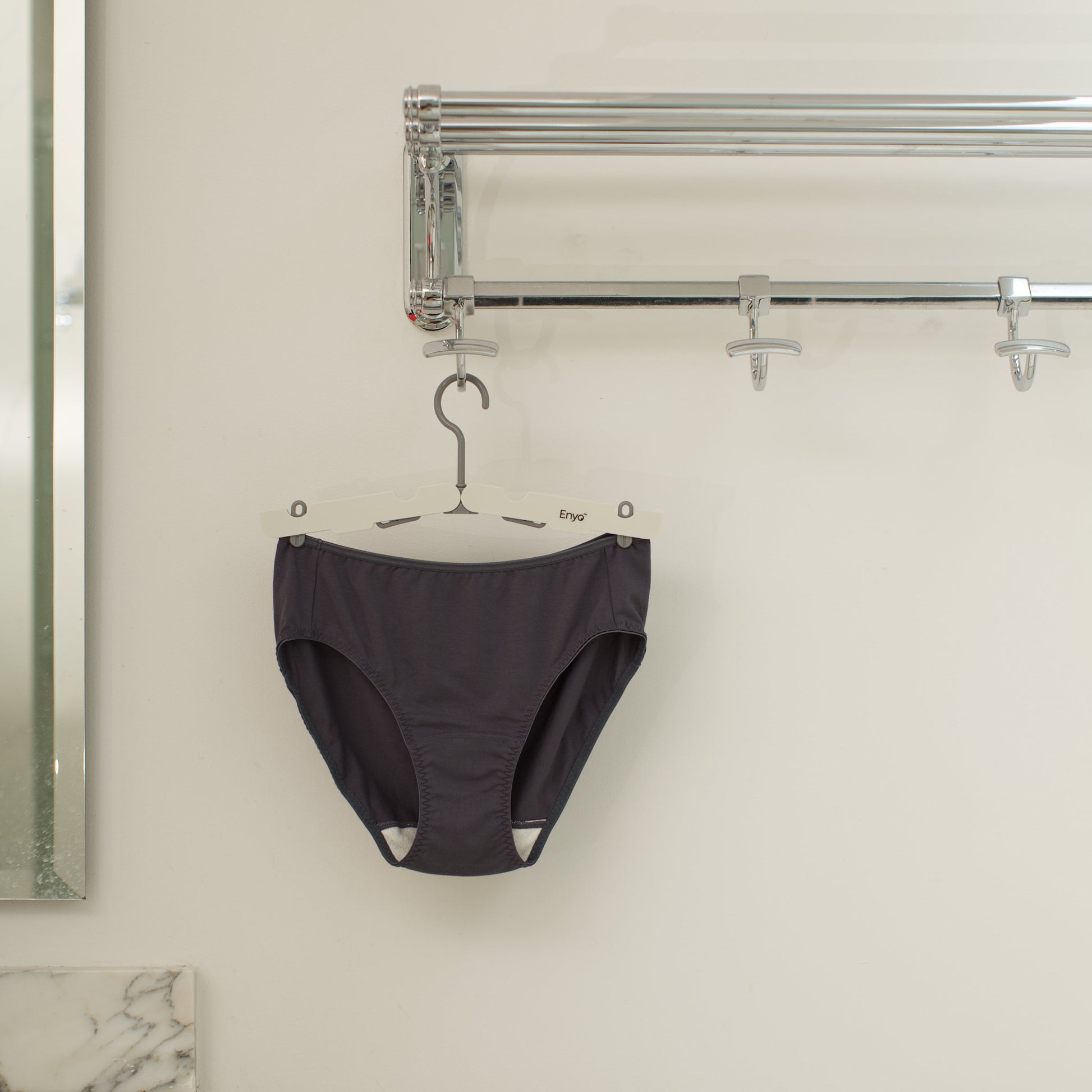 Black underwear with wide white gusset hanging on a 4-clip foldable grey clip hanger on a silver hook against a white wall.