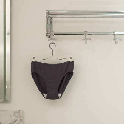 Black underwear with wide white gusset hanging on a 4-clip foldable grey clip hanger on a silver hook against a white wall.