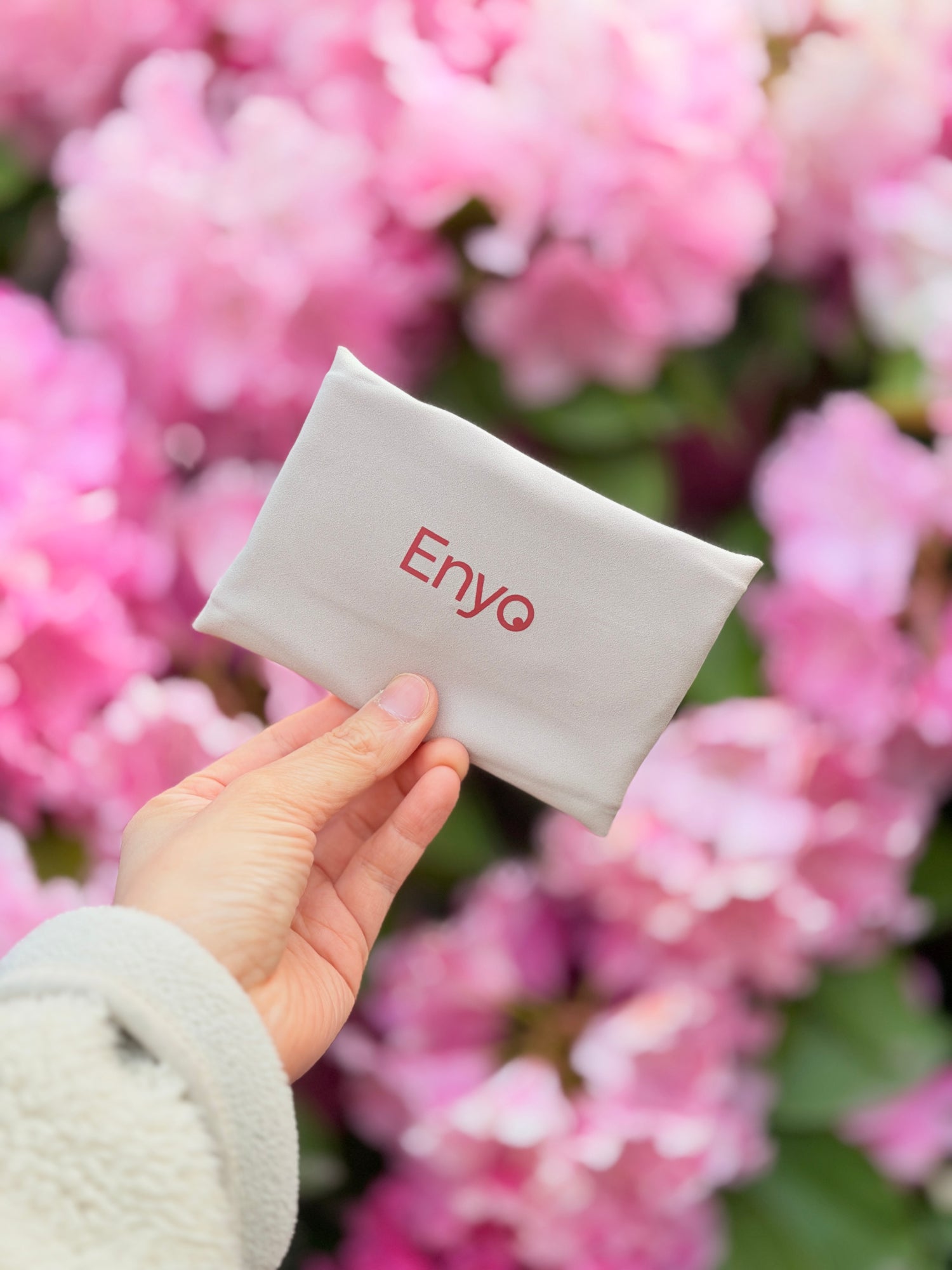 a beige reusable period pad nicely folded in a rectangle with a red text logo, Enyo, on it, being held by a hand in front of pink spring flowers