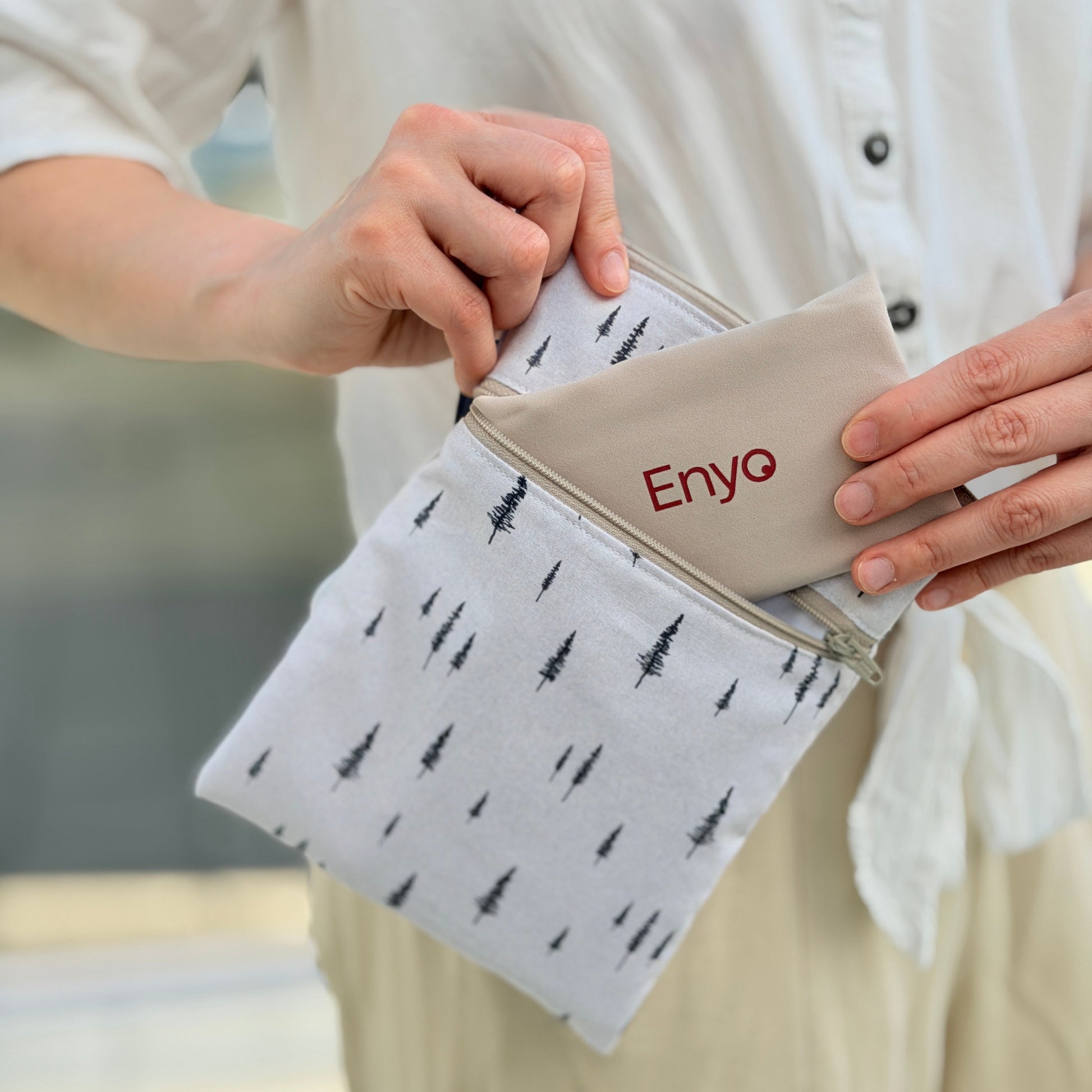 Person holding a folded reusable period pad from a double zipper waterproof wet dry white bag with tree patterns and a beige pouch labeled 'Enyo'.